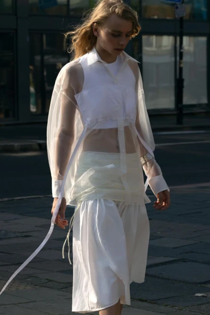 A fair-skinned young adult woman with blond hair walks on a city street wearing a white top, skirt, and transparent shirt. 