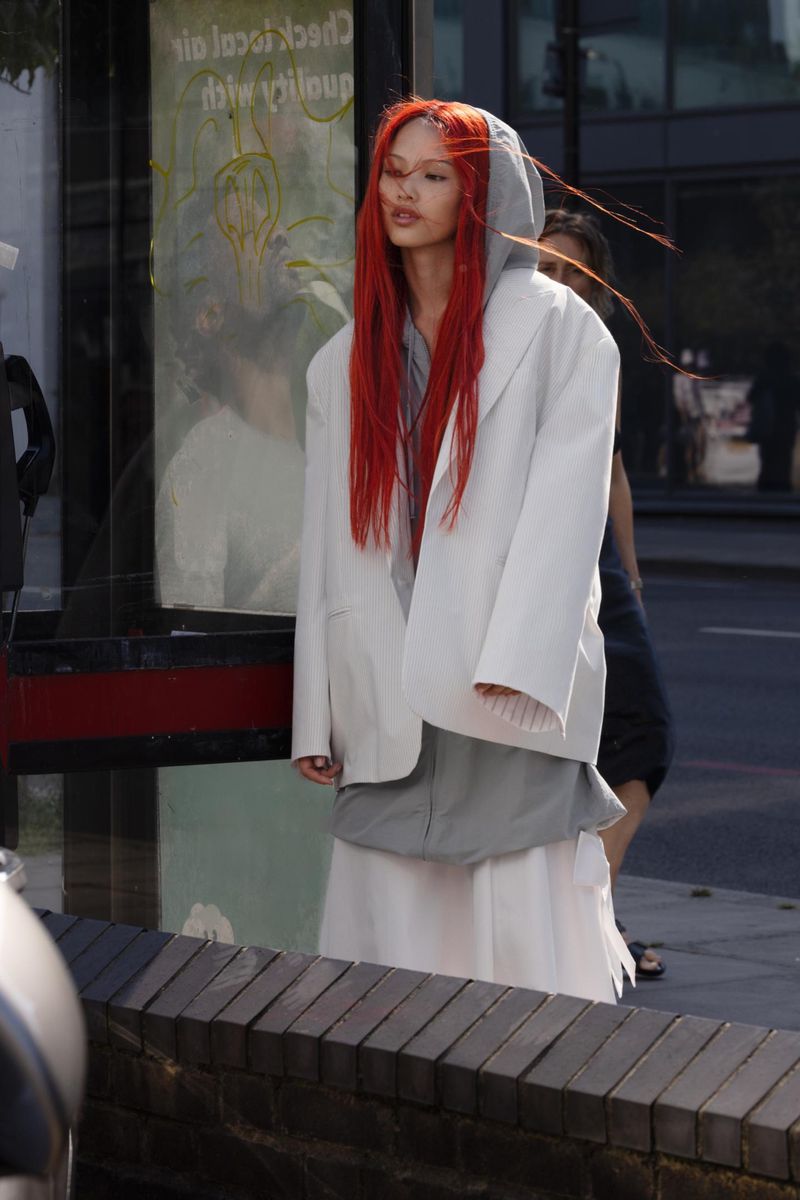 A woman with bright red hair wearing a gray hoodie and a white jacket and skirt, standing on a city street next to a bus stop.