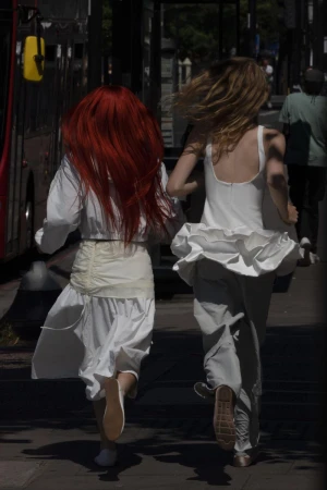 Two young women in white clothing run along a city sidewalk, away from the viewer. One has auburn hair and the other has blonde hair.