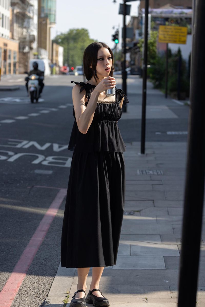 A young adult woman in a black dress stands on a sidewalk holding a can, with a blurred street scene and buildings in the background.