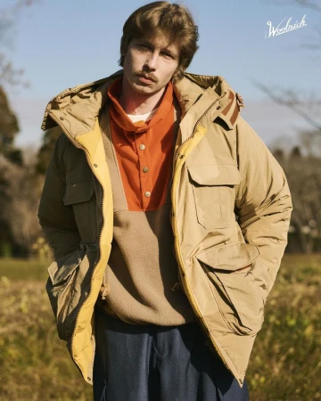 Man in a Woolrich hooded puffer jacket, looking at the camera in an outdoor setting, featuring a light blue sky and fall foliage.