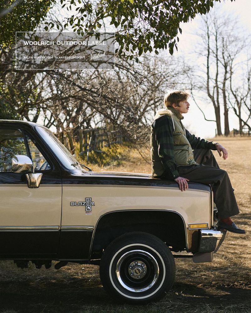 A man wearing a vest and brown pants sits on the hood of a two-tone beige and black pickup truck in a grassy field with trees.