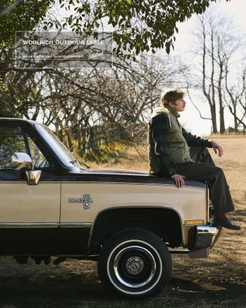 A man wearing a vest and brown pants sits on the hood of a two-tone beige and black pickup truck in a grassy field with trees.