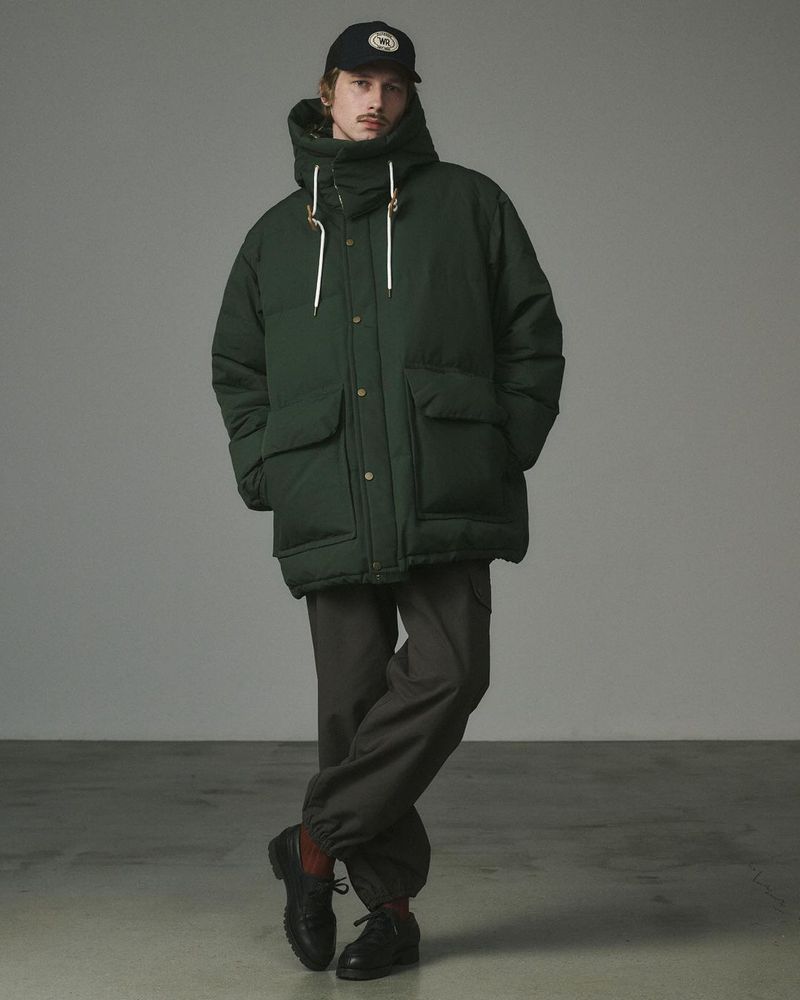 Man in a hooded green puffer jacket, brown pants, and a baseball cap, standing indoors against a neutral backdrop, fashion portrait.