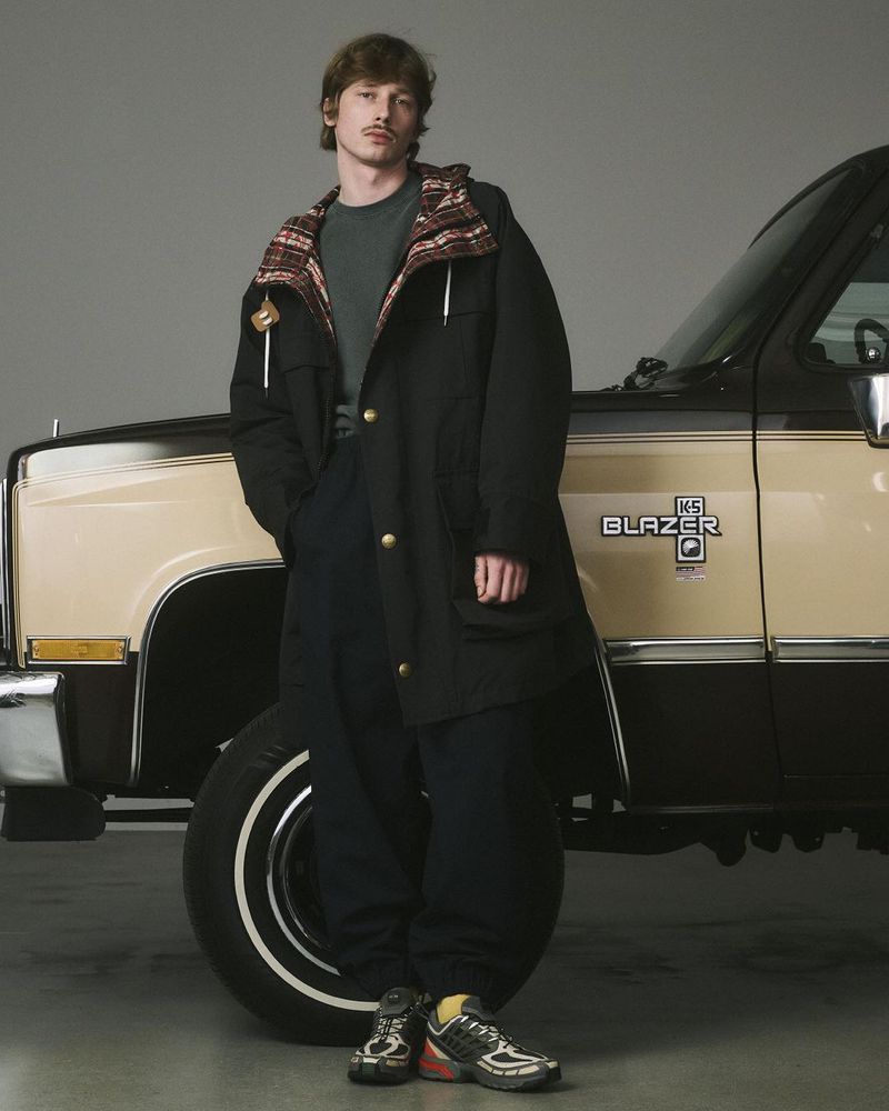A fair-skinned man in a black coat poses next to a beige and black Blazer K5 SUV in a studio, captured in a medium studio shot.