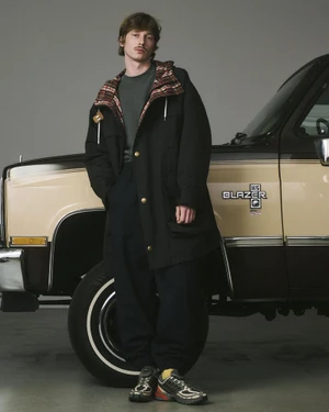 Model wearing dark hooded jacket standing next to a vintage two-tone truck in a studio setting