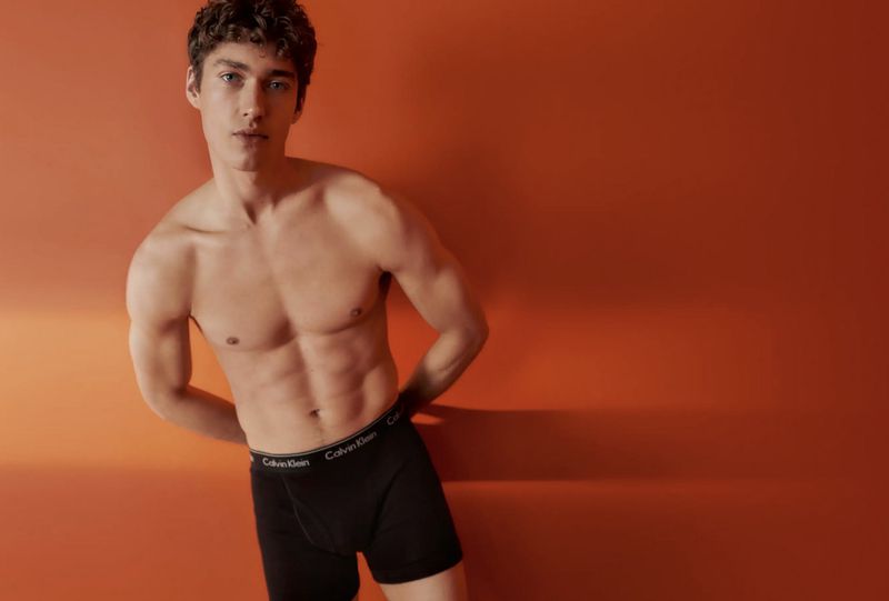 Male fashion model poses against a gradient orange background, showcasing black Calvin Klein underwear in a studio setting, shirtless and toned.