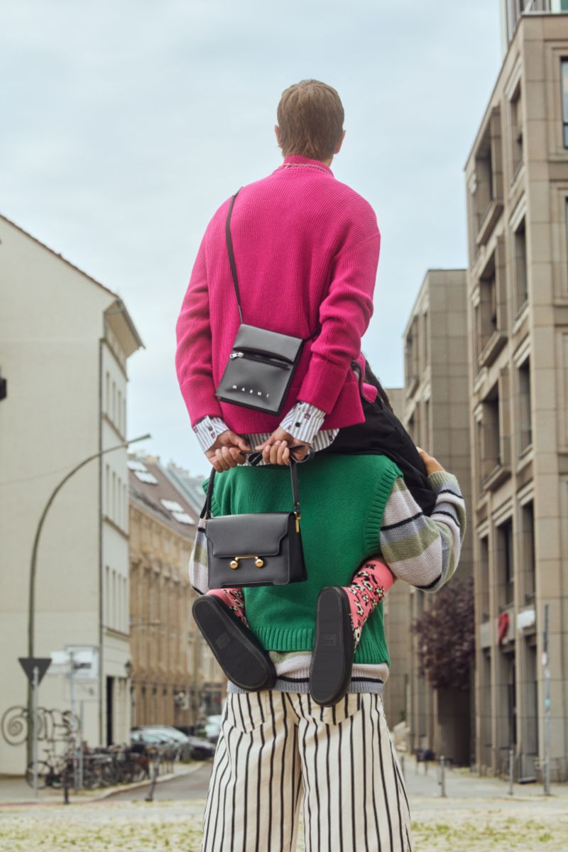 Two people in a city street. One person is carrying another on their shoulders, both with crossbody bags. Buildings and sky in background.