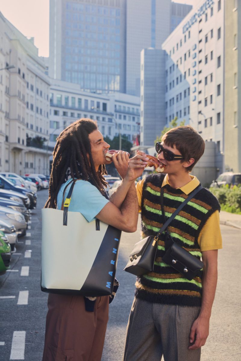 Two men share a sandwich outdoors in an urban setting. One man carries a large tote bag, while the other has a crossbody bag.