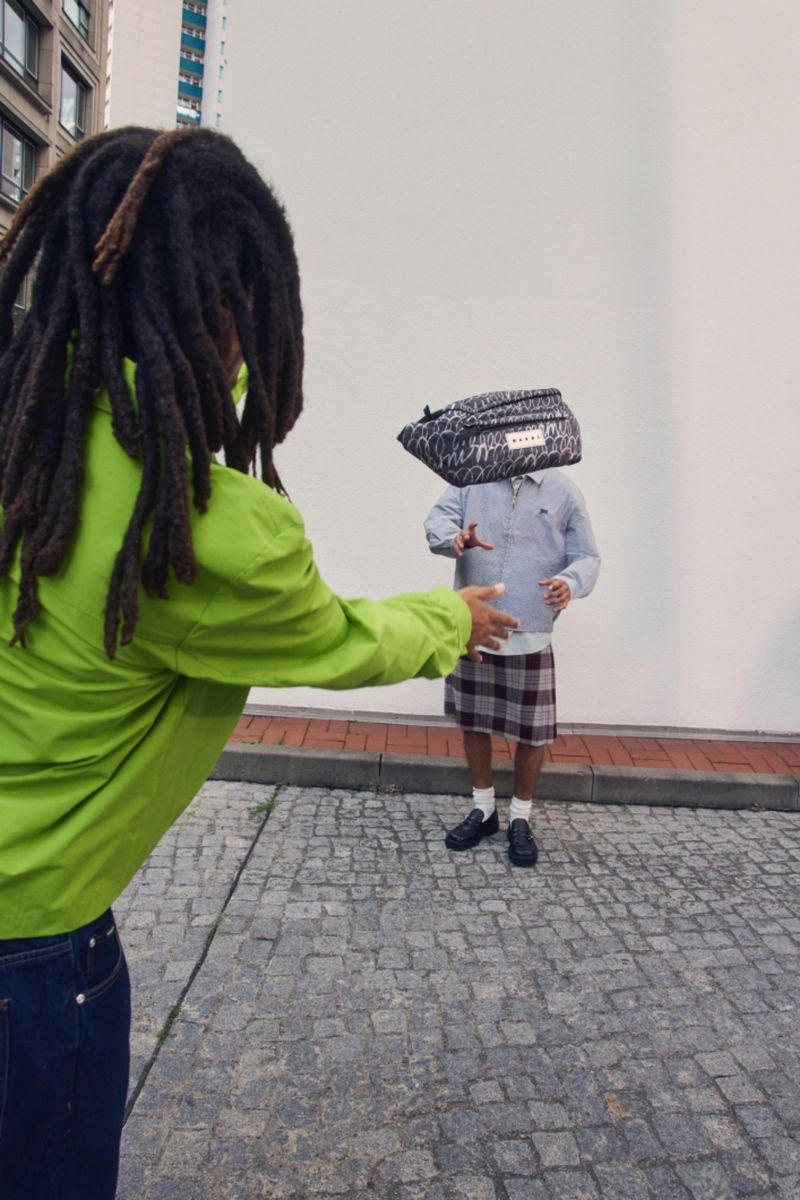 Two people in an outdoor urban setting; one with dreadlocks and a green jacket reaching out to the other who is wearing a checkered skirt.