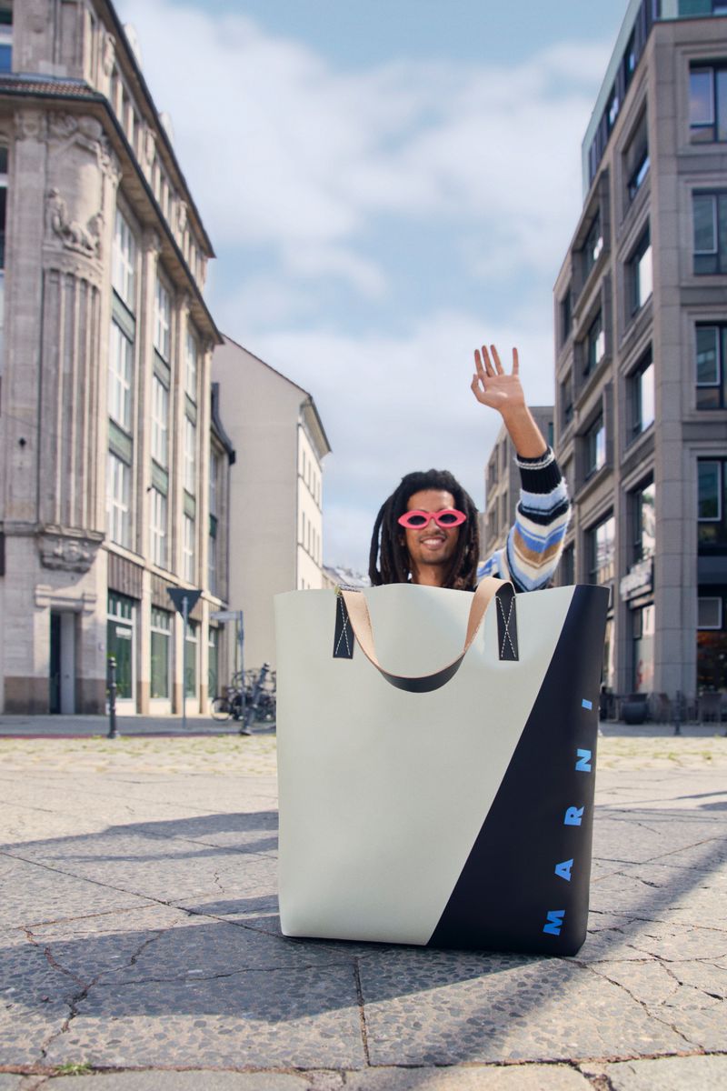 Man stands inside a giant tote bag in an urban street with buildings and cloudy blue sky in the background, fashion shoot.