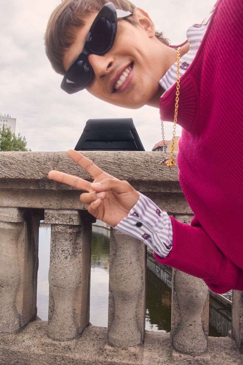 Smiling adult posing on a bridge with a peace sign wearing sunglasses, a pink sweater, and a gold chain; overcast urban setting.