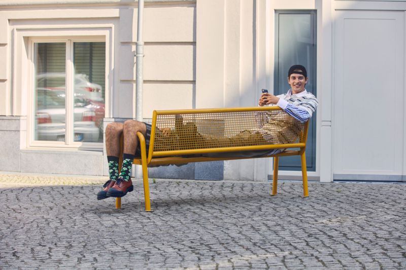 Two young men on a yellow bench on a city street. One man is holding a cell phone, with a light-colored building in the background.