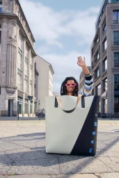 Model in pink sunglasses waving while sitting inside a giant oversized color-block Marni tote bag on a city street.