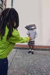 A person with dreadlocks in a bright lime green jacket tosses a patterned black waist bag to a person in a plaid skirt outdoors.
