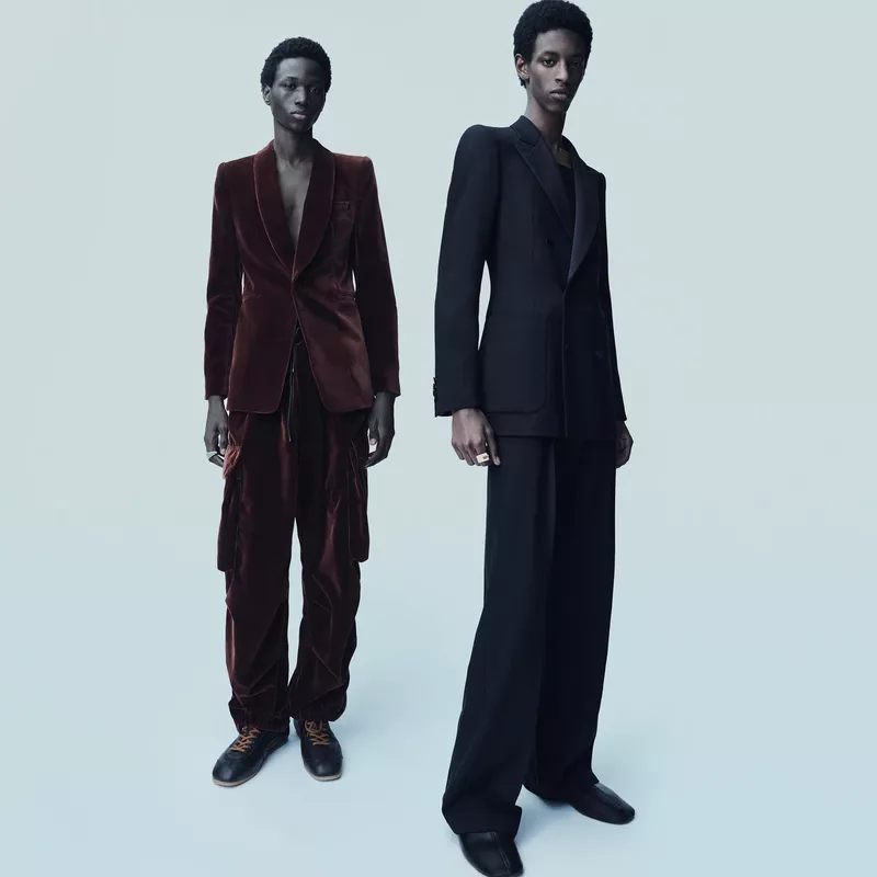 Two Black men in tailored suits pose against a soft blue backdrop; one in a brown velvet suit, the other in a black suit. Fashion editorial.