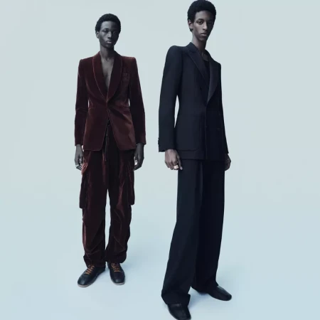 Two Black men in tailored suits pose against a soft blue backdrop; one in a brown velvet suit, the other in a black suit. Fashion editorial.