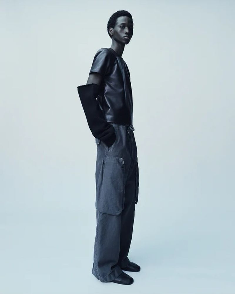 Fashionable young adult man in a dark short-sleeve top, loose grey pants and long gloves posing in a photo studio with a light grey background.
