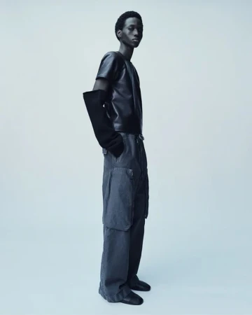 Fashionable young adult man in a dark short-sleeve top, loose grey pants and long gloves posing in a photo studio with a light grey background.