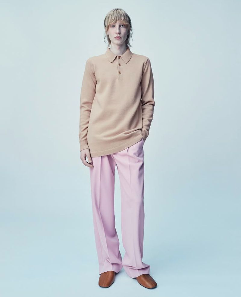 Full-length shot of a male model in a beige polo shirt with long sleeves, pink dress pants, and brown dress shoes, against a studio background.