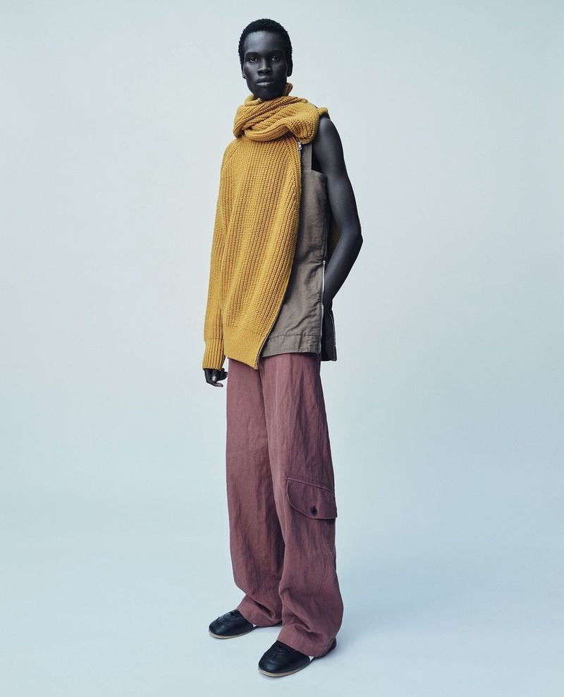 Dark-skinned male model poses in a studio wearing a mustard sweater, earth-toned pants, and black shoes in a fashion editorial shot.