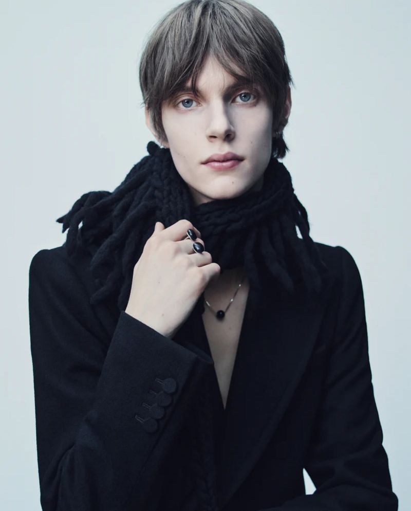 Portrait of a young adult with fair skin, grey hair, and blue eyes wearing a black jacket and textured scarf against a white backdrop.