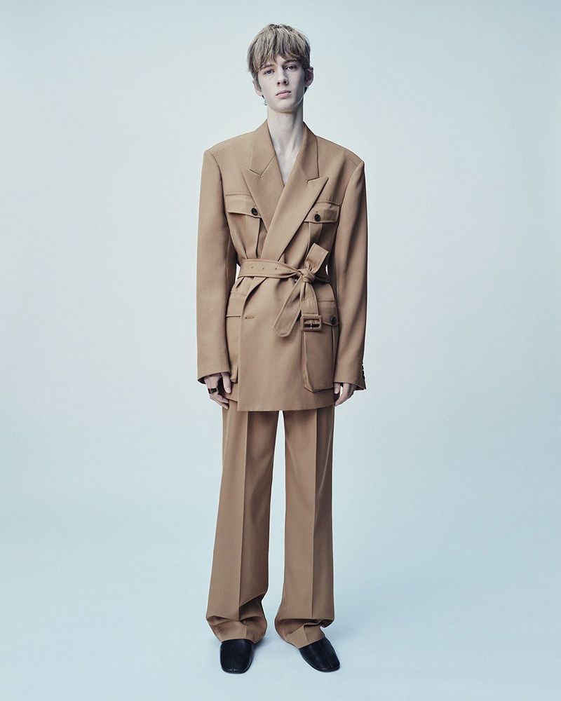 Young adult male fashion model poses in a full-length shot wearing a tan, belted suit with wide-legged pants against a clean backdrop.