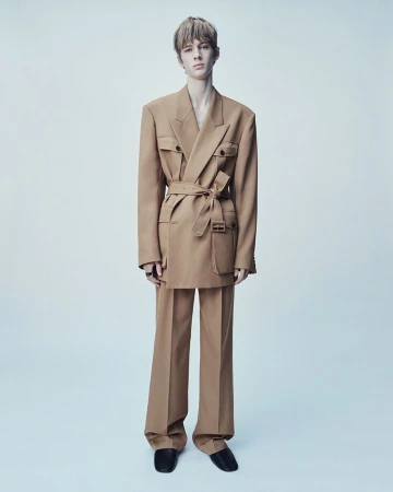 Young adult male fashion model poses in a full-length shot wearing a tan, belted suit with wide-legged pants against a clean backdrop.