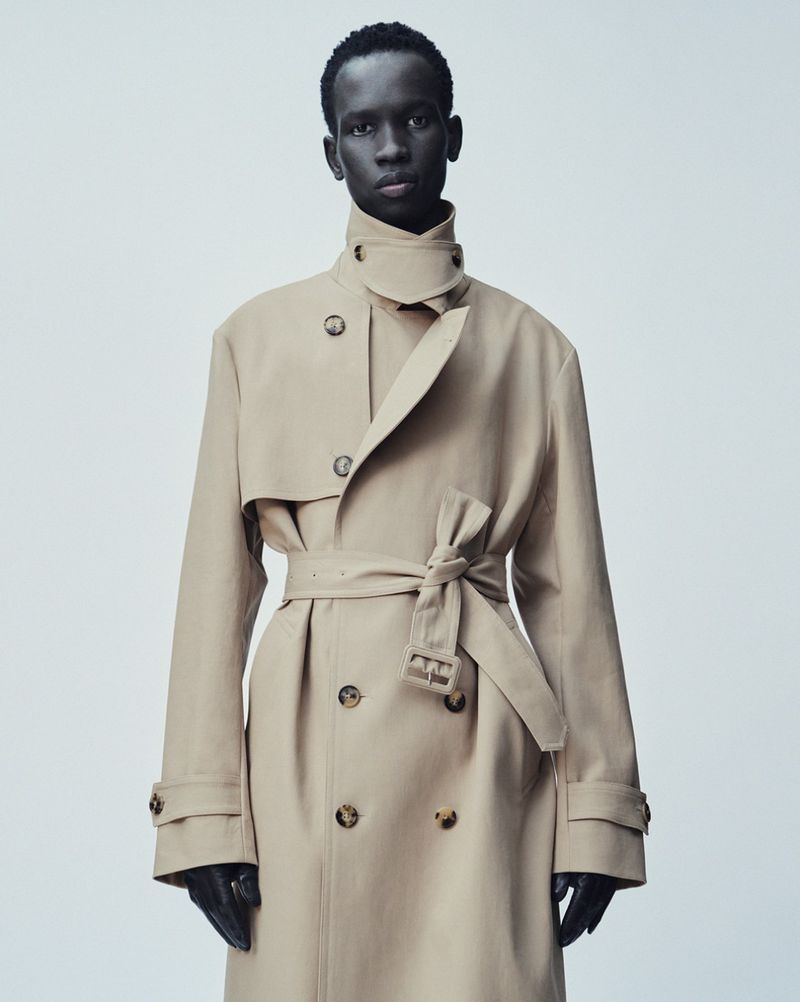 Fashion shot of a single Black male adult in a trench coat. The model is indoors against a plain background, posing for a studio portrait.