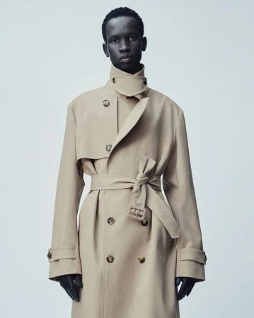 Fashion shot of a single Black male adult in a trench coat. The model is indoors against a plain background, posing for a studio portrait.