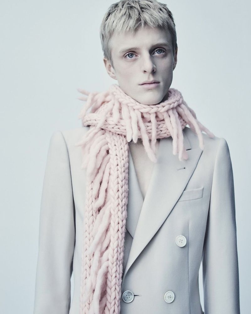 Blond male fashion model in a light grey suit and pink fringe scarf. Fashionable clothing in a studio setting for editorial use.