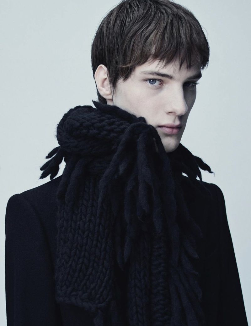 Male model wearing a chunky black knit fringed scarf and tailored coat against a pale studio backdrop.
