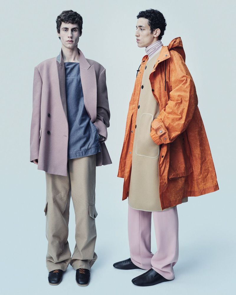 Two young men showcase modern menswear fashion, one in a pink blazer with tan pants, the other in an orange raincoat with pink trousers.