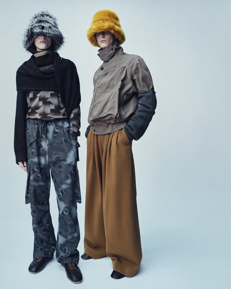 Two models in avant-garde fashion featuring oversized trousers, unique tops, and furry hats against a bright, neutral backdrop. 