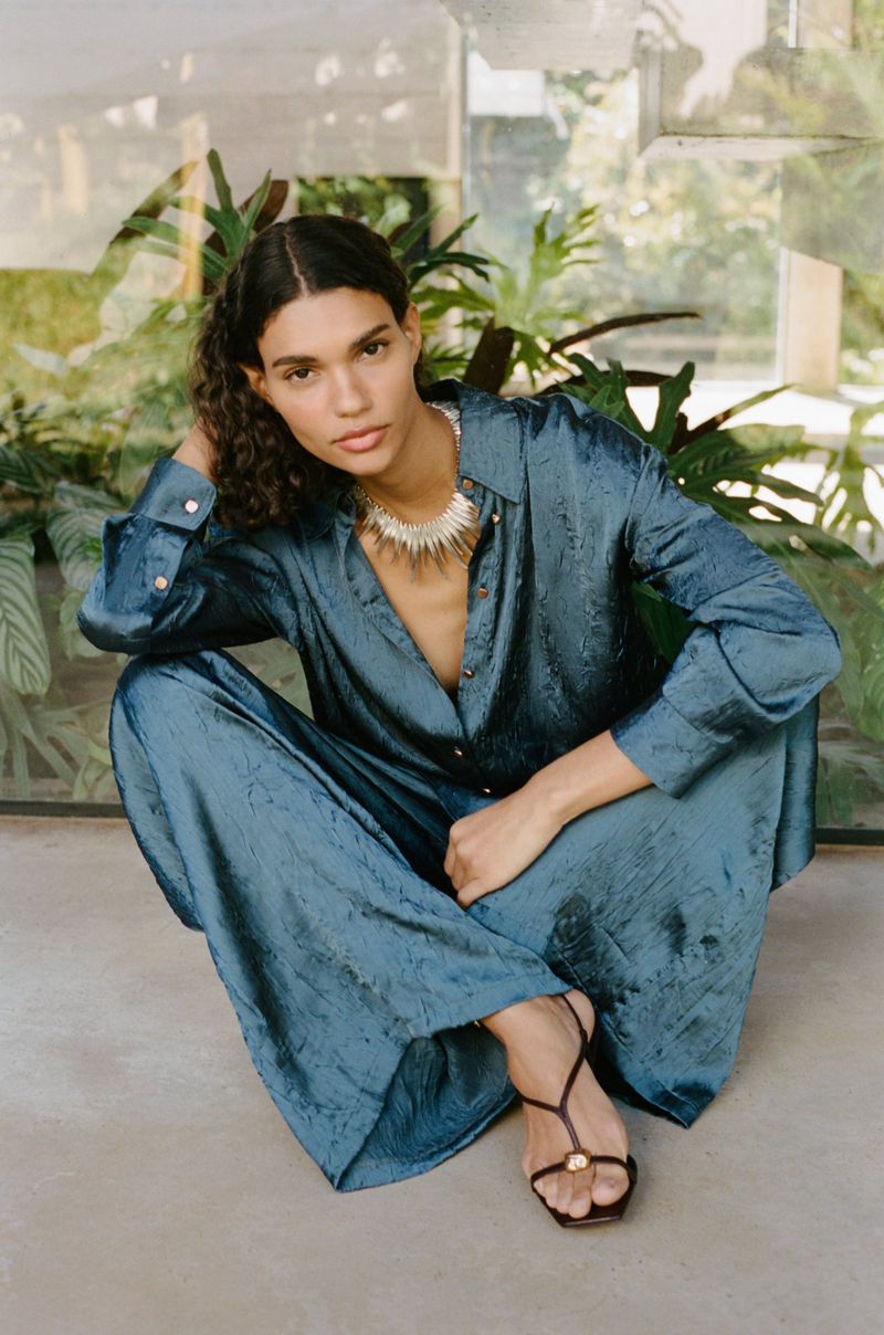 Elegant woman poses in a matching teal satin shirt and palazzo pants, accessorized with strappy sandals and a statement necklace, sitting indoors near foliage.