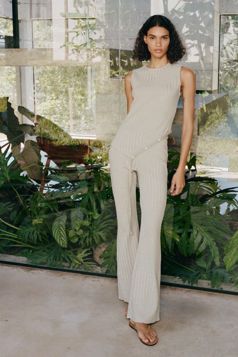 An adult woman poses indoors wearing a beige ribbed sleeveless top and matching pants, standing in front of a window with green foliage.