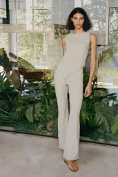 Model Barbara Valente wearing a beige sleeveless ribbed knit set in front of glass windows overlooking lush greenery.