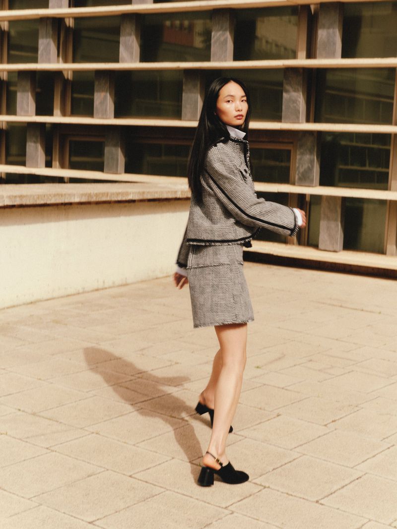 A woman in a grey tweed skirt suit and black heeled shoes poses in an outdoor setting with an architectural background on a sunny day.