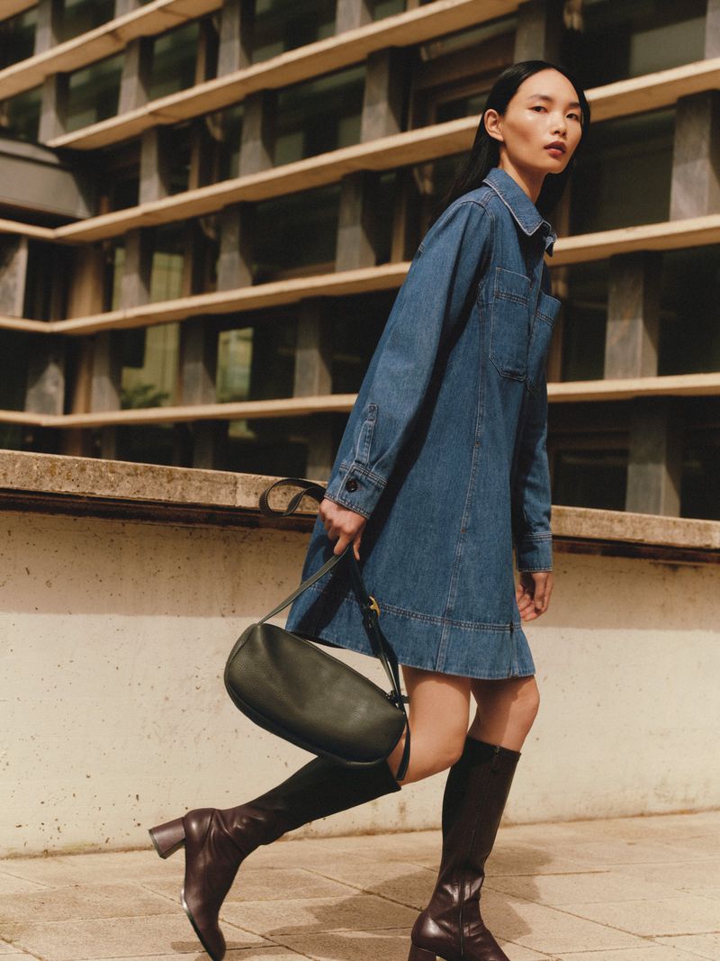 An Asian woman models a blue denim dress and high brown boots, carrying a black purse, posing outside a building in daylight.