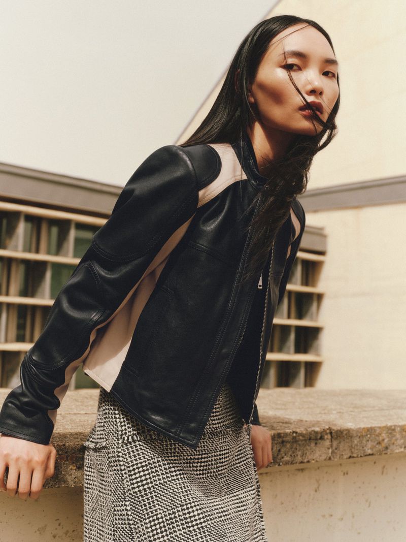 An Asian fashion model poses outdoors wearing a black leather jacket and a houndstooth skirt, leaning against a wall.