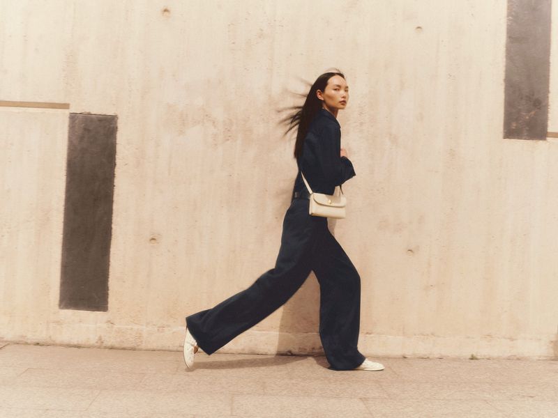 A stylish Asian woman walks in a navy suit and cream purse against a minimalist beige wall, exuding modern fashion and urban confidence.