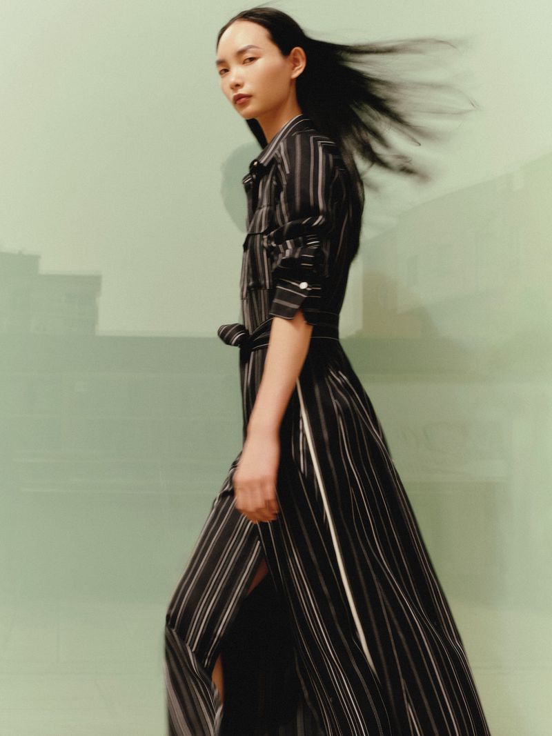An Asian woman walks and looks over her shoulder, wearing a striped black and white dress with flowing black hair against a light green background.