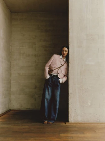 Fashion shot of a young adult Asian woman in wide-leg jeans and a pink tweed jacket, standing against a concrete wall in a neutral interior.