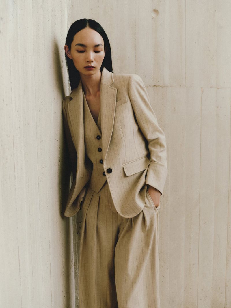 Adult female wearing a beige pin-striped suit with a matching vest, leaning against a beige wall in a studio setting.