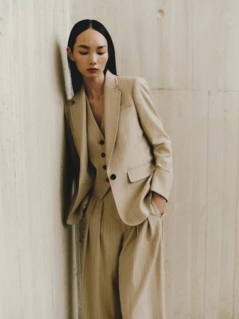 Adult female wearing a beige pin-striped suit with a matching vest, leaning against a beige wall in a studio setting.