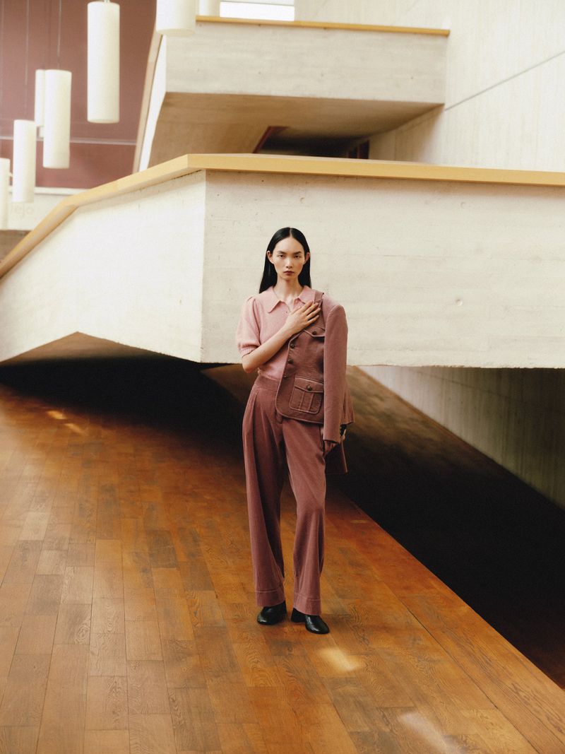 Fashion shot of an Asian woman in a mauve suit standing on a wood floor in a concrete building with pendant lights overhead.
