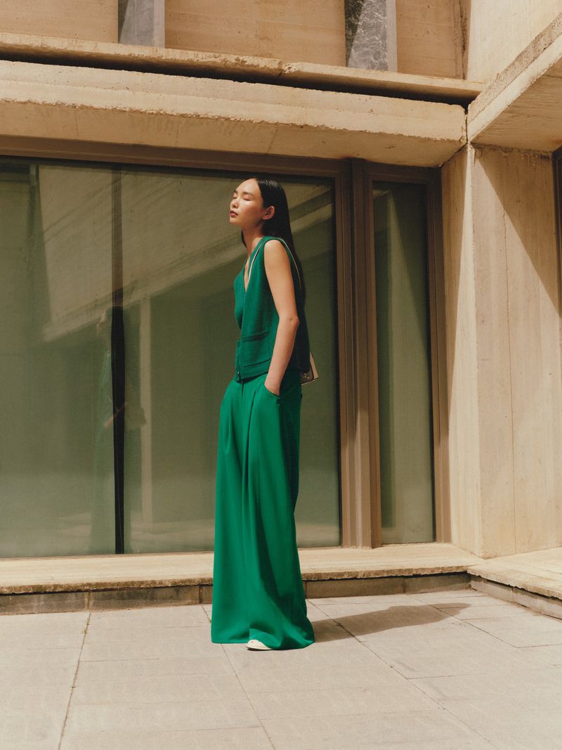 A woman in green wide-leg pants and a sleeveless top poses against a tan concrete building with windows, in a fashion editorial style.