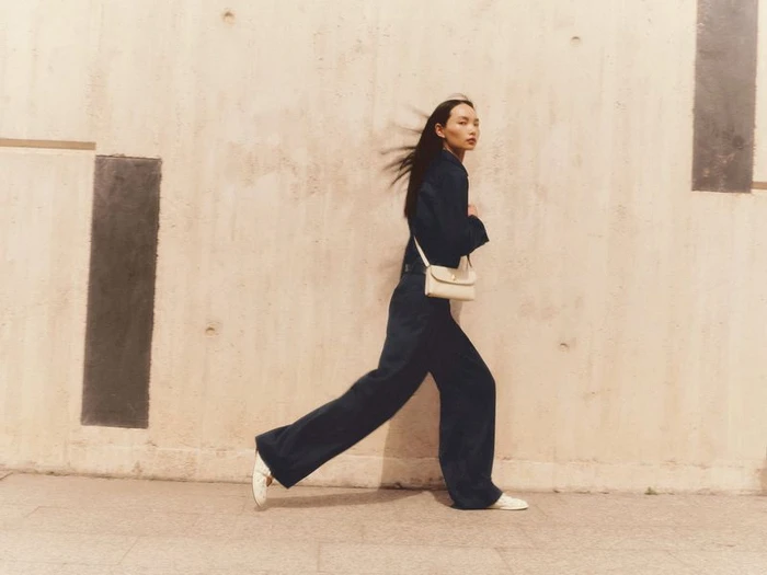 Model Ling Chen strides in a dark navy jumpsuit with a cream crossbody bag against a warm concrete wall for ME+EM.