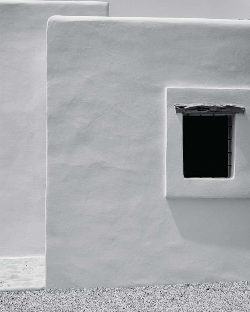 Minimalist architectural detail featuring a white adobe building with a small window casting a sharp shadow on a gravel surface.