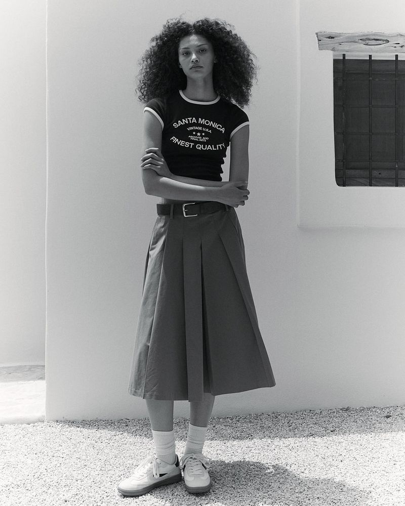Full shot of a young, slim adult female with curly hair in a dark tee and long pleated skirt, posing outdoors in a minimalist style.